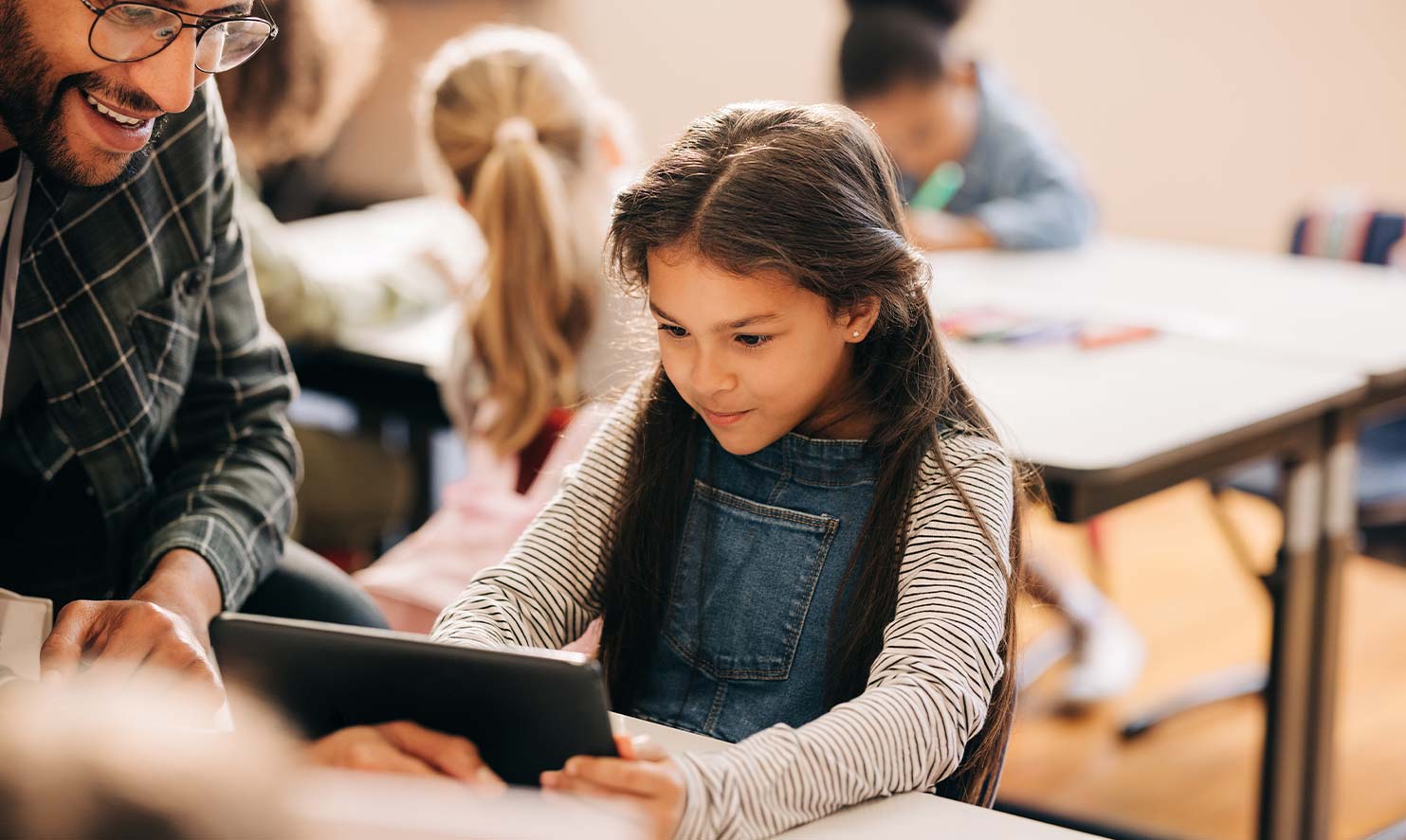 Ein junges Mädchen arbeitet im Unterricht mit einem Tablet. Ein Lehrer schaut ihr über die Schulter.